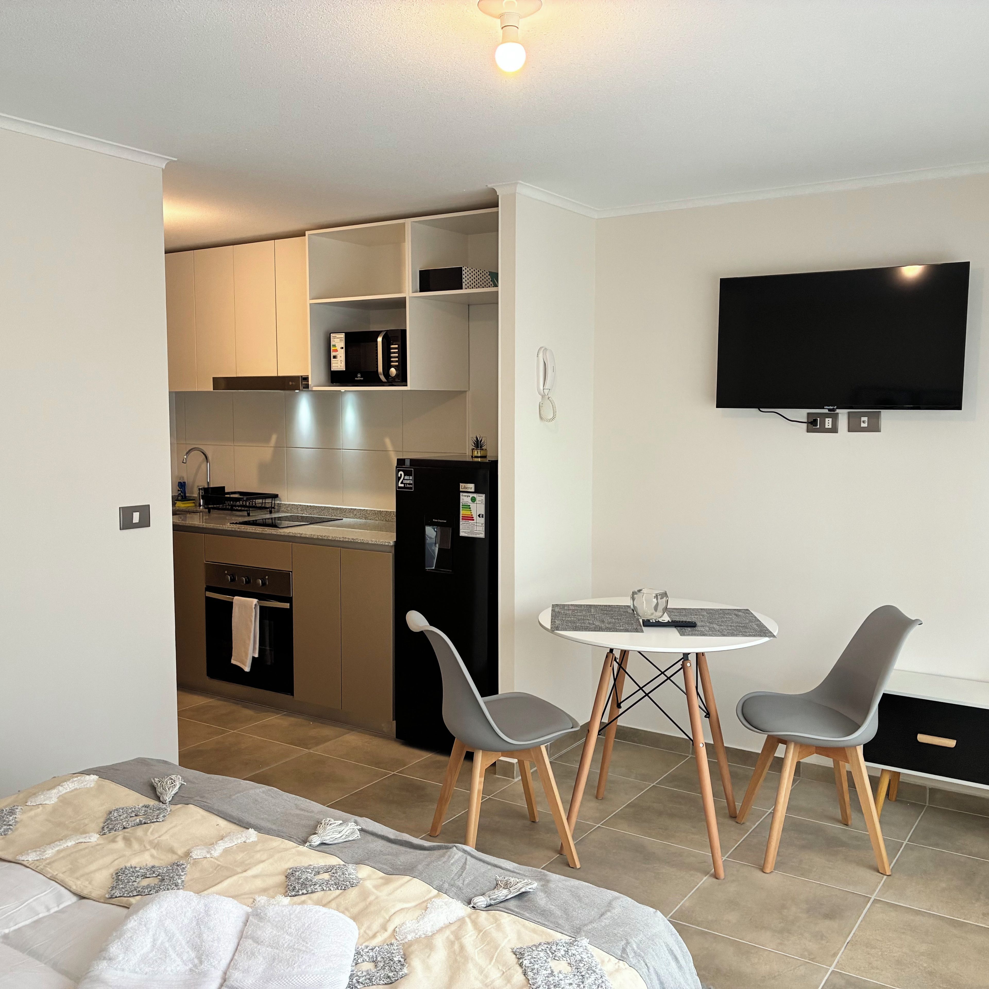 A compact hotel room with a kitchenette, a dining area with two chairs, a bed, and a flat-screen TV mounted on the wall.