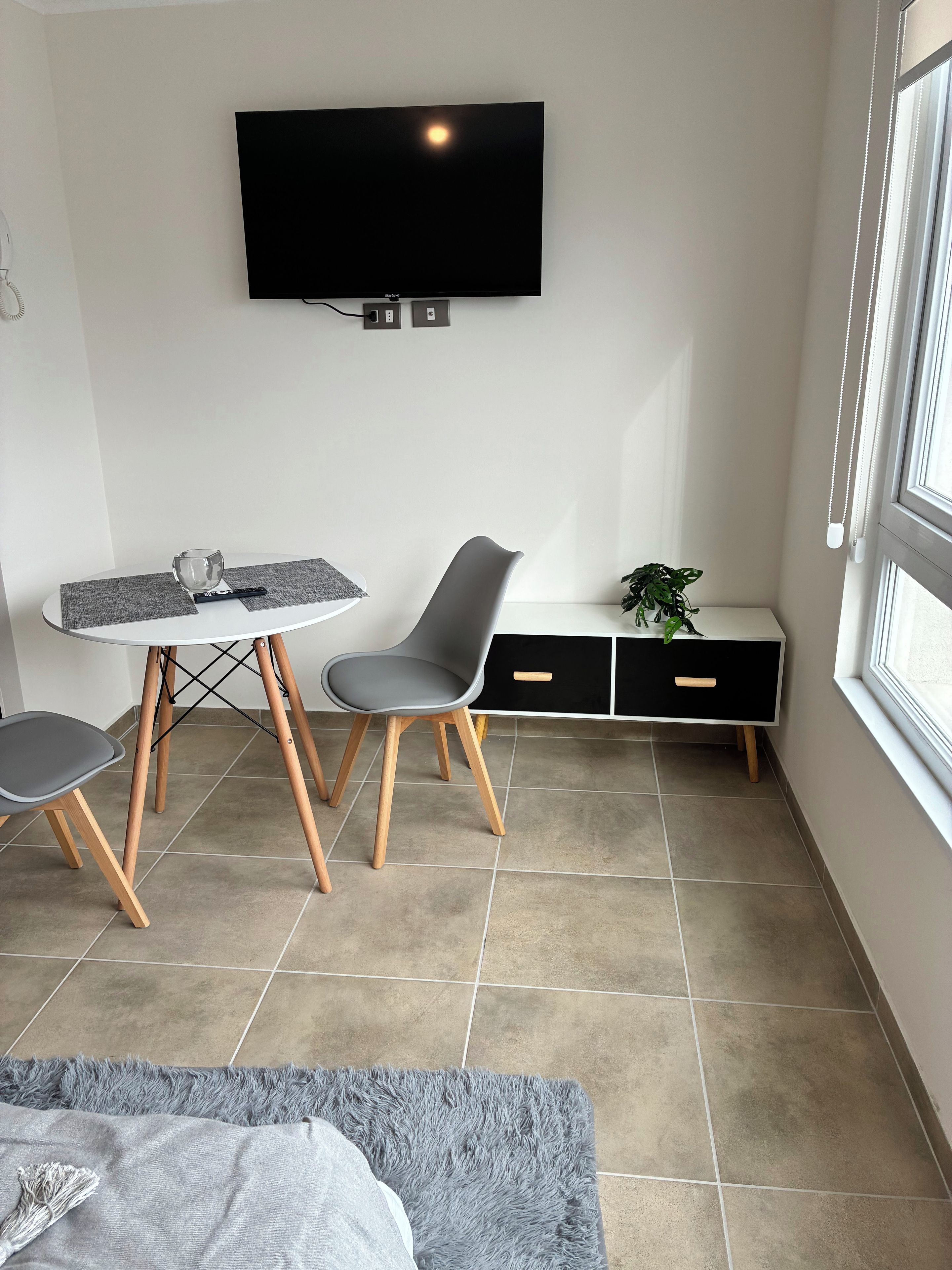 A modern, compact living space with a flat-screen TV mounted on the wall, a dining table with chairs, a bench with storage, and a large window providing natural light.