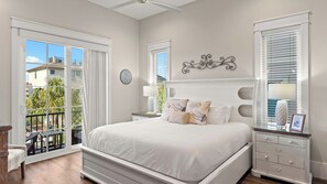 Een slaapkamer met een groot bed, een nachtkastje, een dressoir en een balkon met uitzicht op gebouwen en groen.