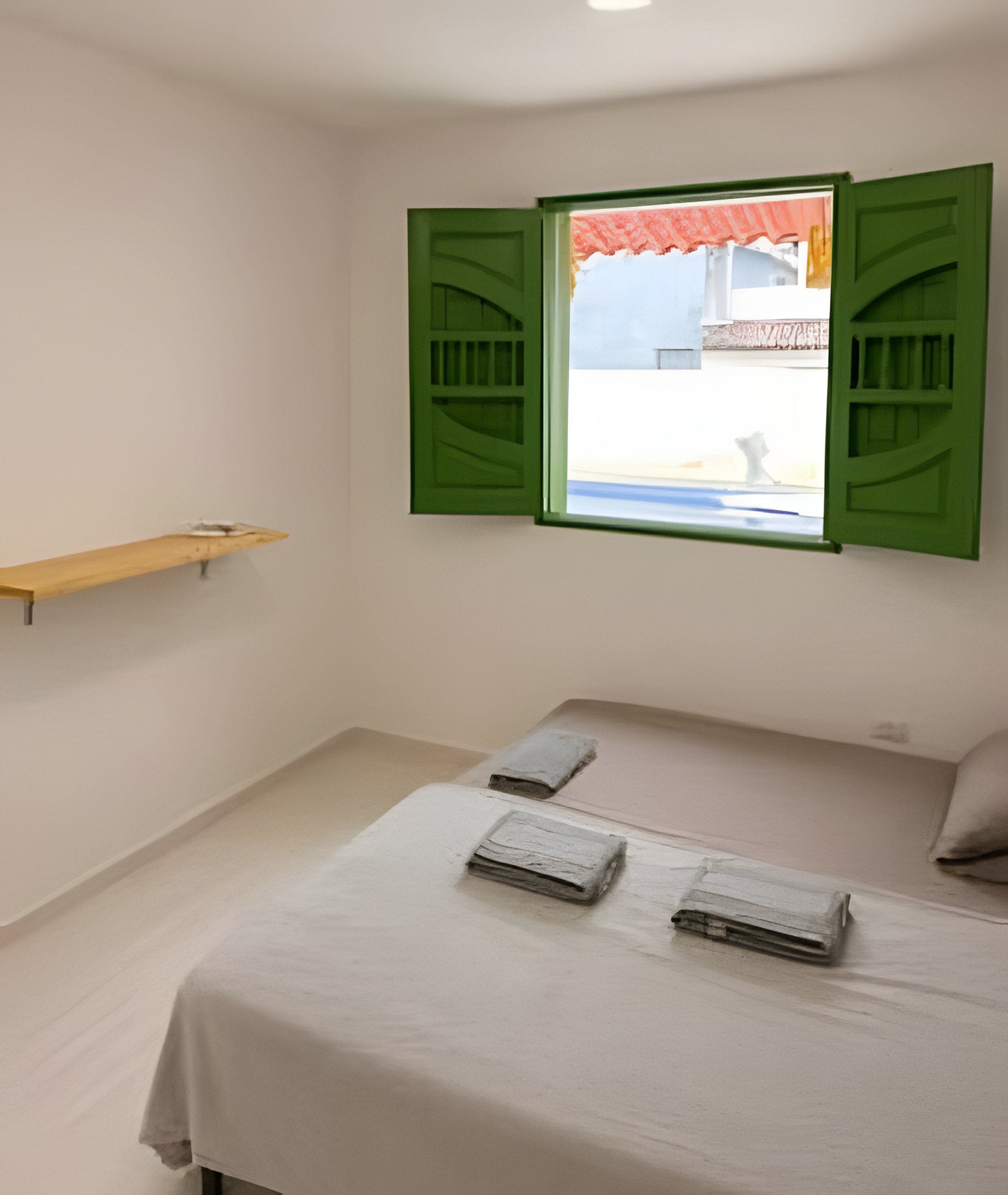 Un dormitorio con cama, dos almohadas, estante de madera y ventana con contraventanas verdes.