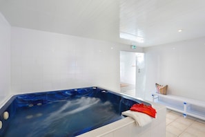 A modern bathroom with a large blue hot tub, white tiled walls, and a bench with towels. - T3 avec vue sur les montagnes | Piscine intérieure (Germ)