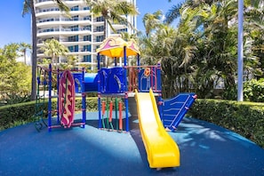 Ein Spielplatz mit Rutsche und Klettergerüsten, umgeben von Palmen und einem Gebäude im Hintergrund.