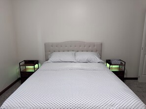 Un llit amb un panell de capçal guarnit, dues taules de nit amb làmpades i un edredó estampat.