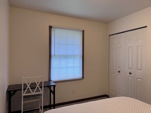 Ein Schlafzimmer mit einem Bett, einem Schreibtisch und einem Stuhl. Es gibt ein Fenster mit Jalousien und eine Reihe weißer Türen.