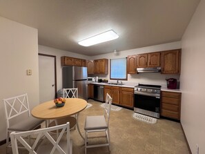A compact kitchen with wooden cabinets, a stainless steel refrigerator, a microwave, a stove, and a sink. There is a dining table with chairs and a bowl of fruit. - Betty's in Berrien Springs Near AU Newly Renovated (Berrien Springs)