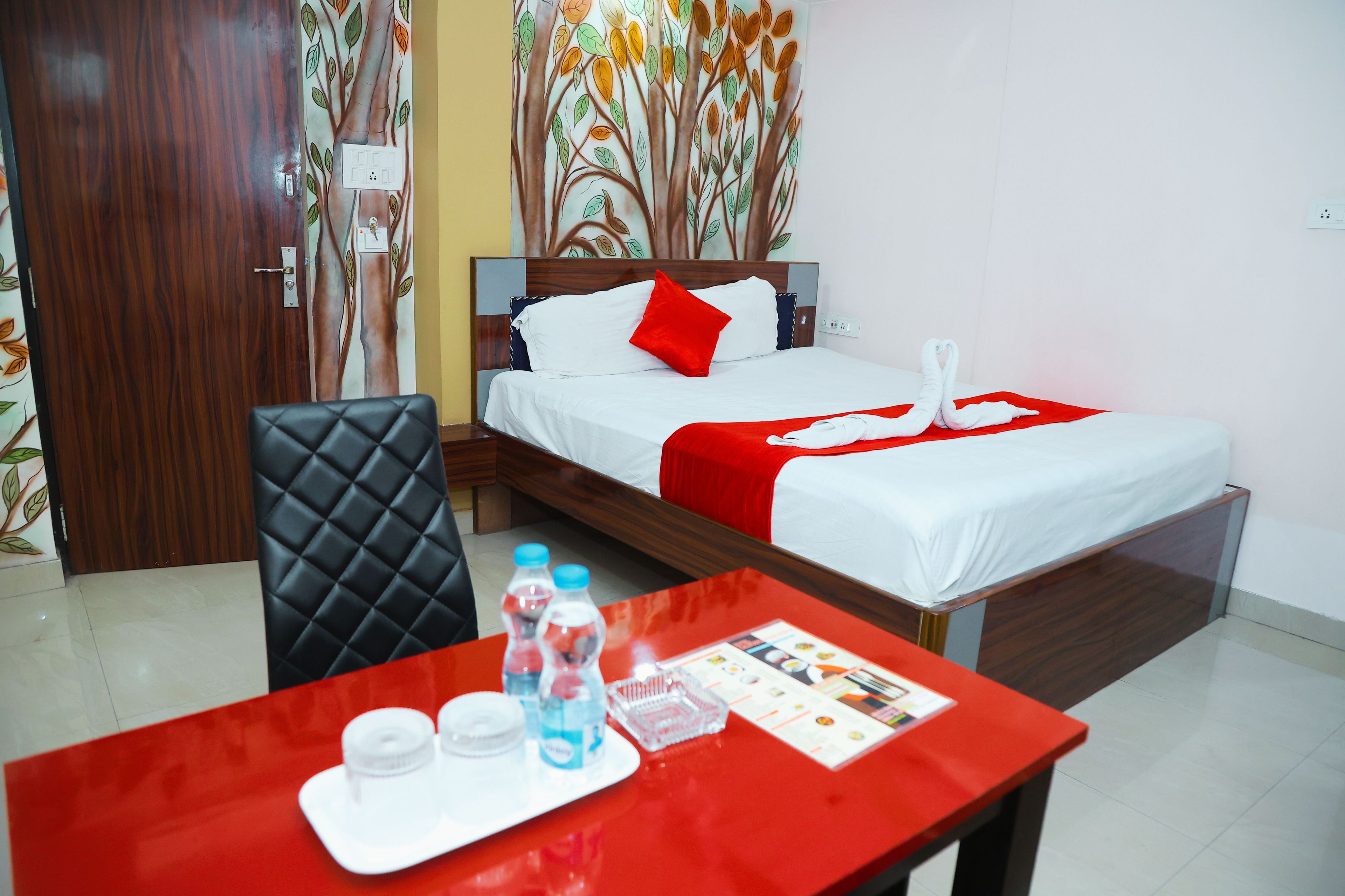 A hotel room with a bed, a desk with a red table, a chair, and a wall with a tree pattern.