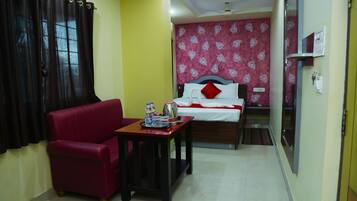 A compact hotel room with a red sofa, a small table with a tray of items, a bed with white bedding and a red pillow, and a patterned wall.