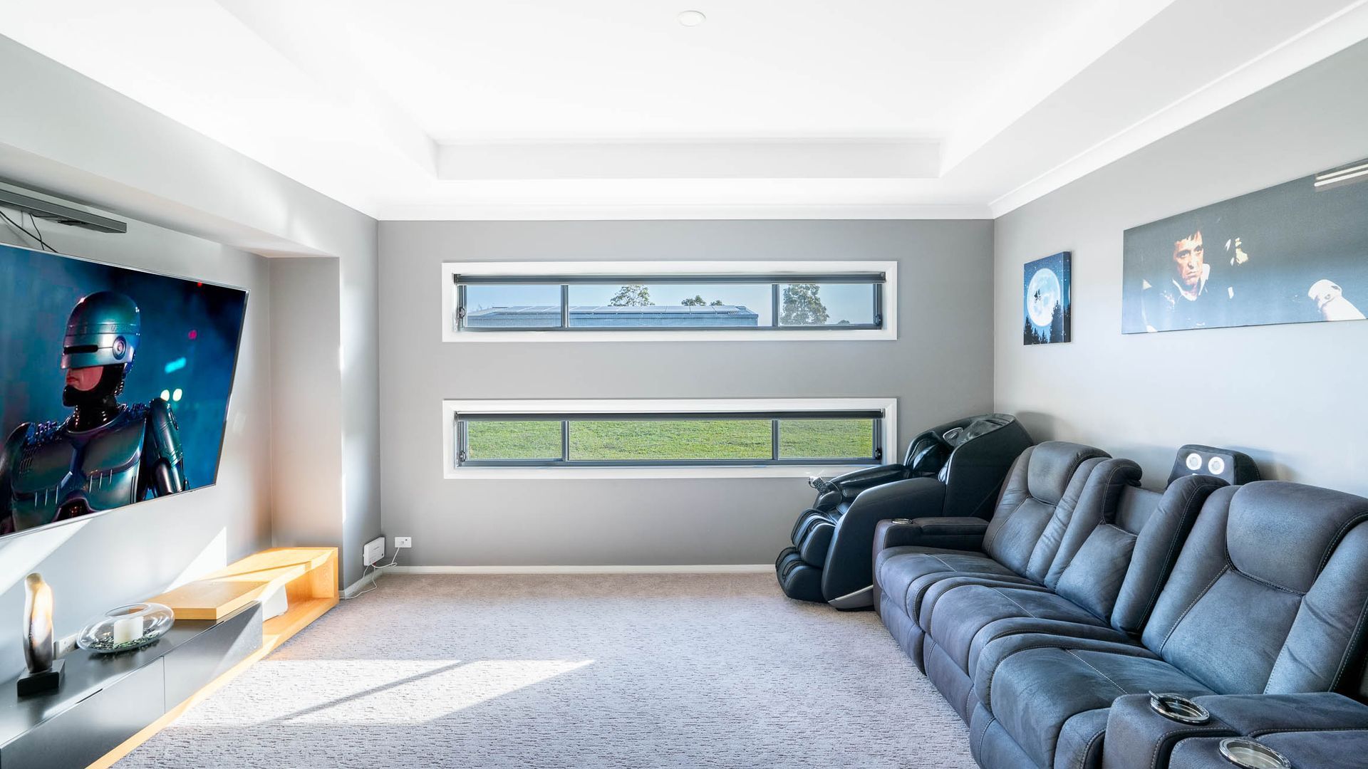 Un salon moderne doté d’un grand téléviseur à écran plat, d’un canapé gris et de deux fenêtres donnant sur un immeuble et sur la verdure.