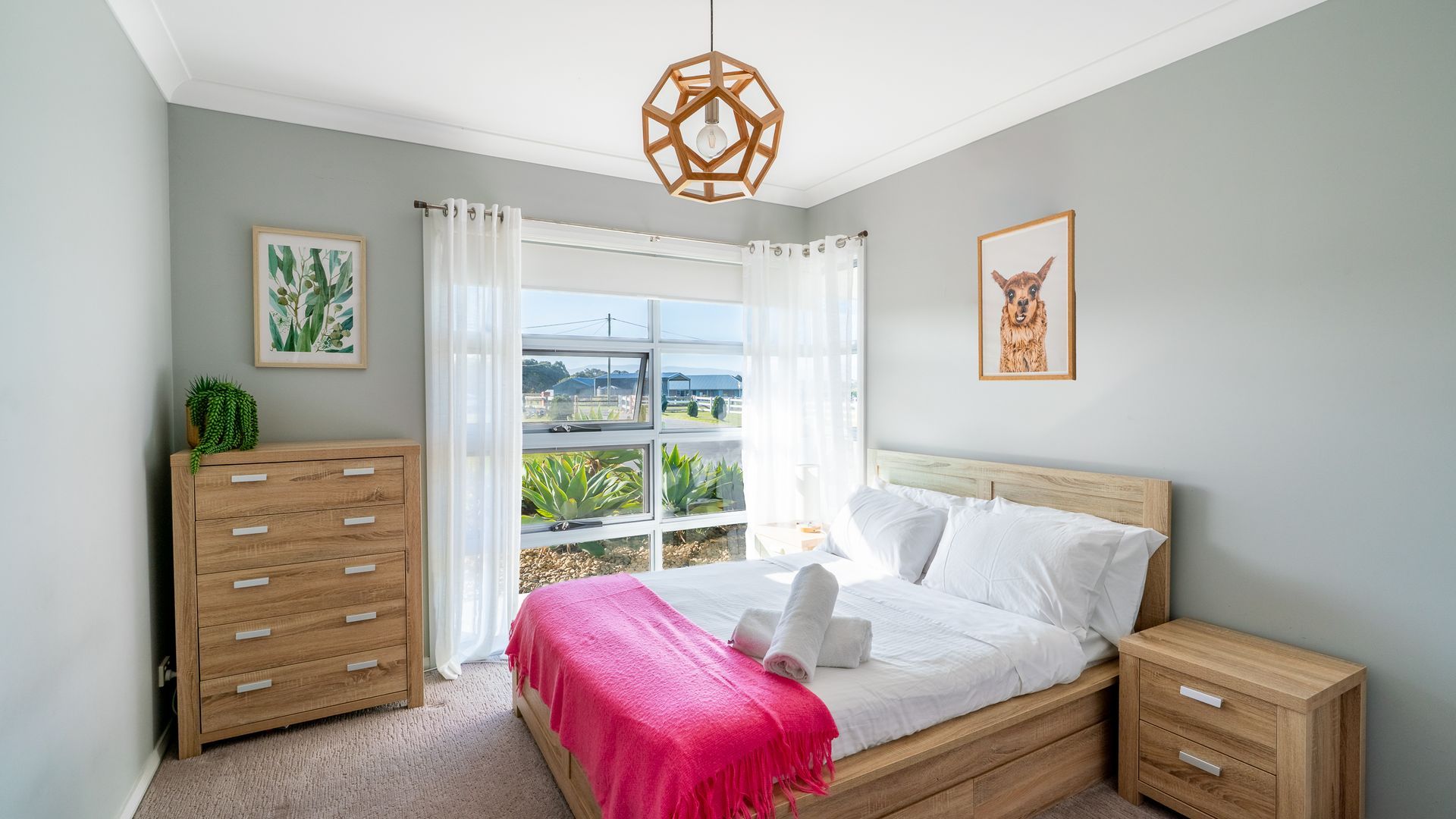 Une chambre à coucher comprenant un lit, une table de chevet, une commode, une fenêtre avec des rideaux et un luminaire suspendu.