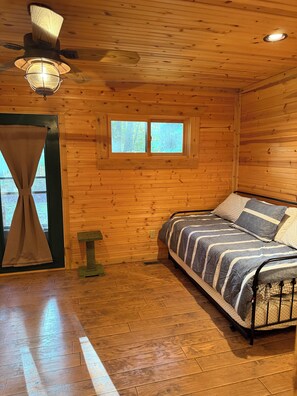 Ein Zimmer in einer Holzhütte mit einem Bett, einem Fenster, einem Deckenventilator und einem Nachttisch.