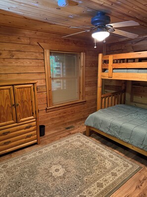 A wooden cabin interior with a bunk bed, a cabinet, a window with blinds, and a ceiling fan. - 4-bedroom log cabin in Gladwin on the Tittawassee River with its own boat launch (Gladwin)