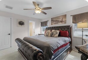 Quarto com cama grande, ventilador de teto, janela com persianas e uma obra de arte na parede.