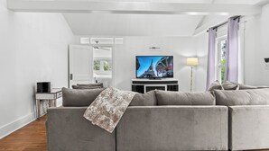 A modern living room with a grey sectional sofa, a flat-screen TV, a floor lamp, and a large window with curtains. - StayHaus - The Signature Flat (St. Petersburg)