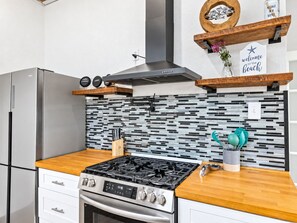 A modern kitchen with a stainless steel refrigerator, a gas stove, and a microwave. There are wooden shelves with decorative items and a sign that reads 'welcome to the beach.' - Pet-Friendly 2BR Galveston Retreat | Cowboy Pool | Sleeps 8 (Galveston)