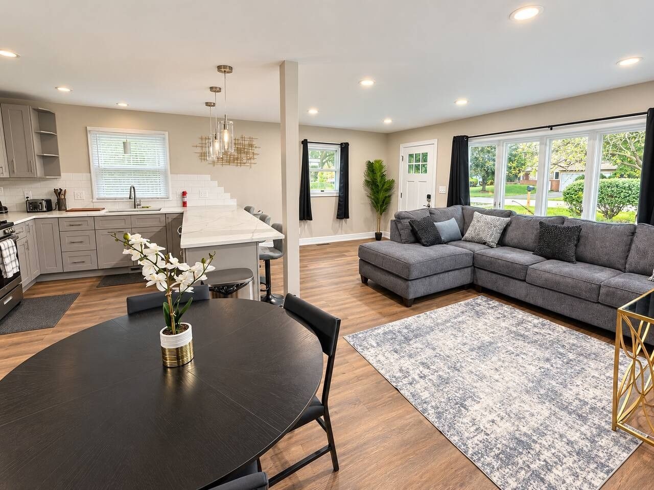 A modern kitchen with a dining area, a gray sectional sofa, and a large window with a view of greenery.