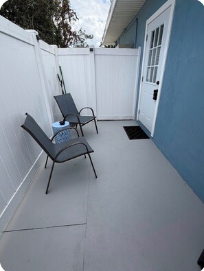 A covered patio area with two metal chairs and a small table, a white fence, and a blue door. - Cozy 1 bedroom studio in lovely Brandon with AC (Brandon)