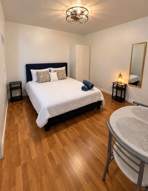 Quarto com cama, mesas de cabeceira, uma mesa redonda com cadeira, luminária no teto e espelho na parede.