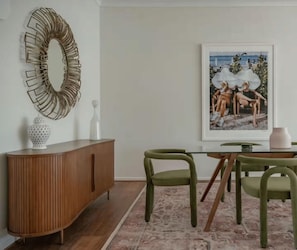 A dining area with a wooden cabinet, green chairs, a glass table, and a framed artwork on the wall. - Lõco Píso House Pet friendly - Pool & Spa (Brisbane)