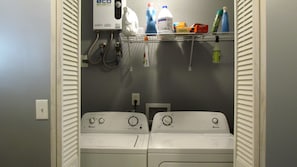 A laundry room with two front-loading washing machines, a water heater, and shelves with cleaning supplies. - Downstairs carriage house on Columbus (Savannah)
