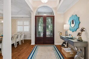 A well-lit entryway with a wooden door, a dining area with white chairs, and a decorative wall piece. - Palmetto Dunes Estate for 20, Heated Pool, EV Charger, 9 Min to Beach (Hilton Head Island)