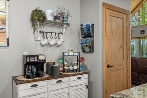Une cuisine équipée d’une machine à café, de tasses et d’une étagère où sont posées des plantes et des magazines.