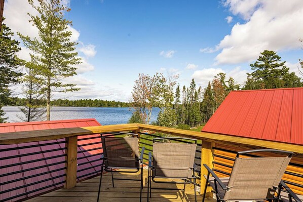 Une terrasse avec des chaises donnant sur un lac, entourée d’arbres et bordée d’un bâtiment au toit rouge.