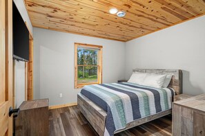 Une chambre avec un plafond en bois, un lit recouvert d’une couverture à rayures, une fenêtre donnant sur des arbres et une table de chevet en bois.