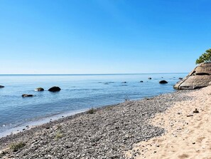 Ein kiesiger Strand mit klarem, blauem Wasser und einem Felsen im rechten Bildrand.
