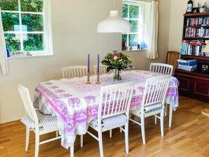 Comedor con mesa dispuesta para seis personas, rodeada de sillas blancas, una lámpara colgante blanca y una estantería al fondo.