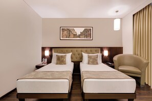 Ein Hotelzimmer mit zwei Betten, einem Kopfteil, Nachttischen, einer Lampe, einem Sessel und einem gerahmten Bild an der Wand.