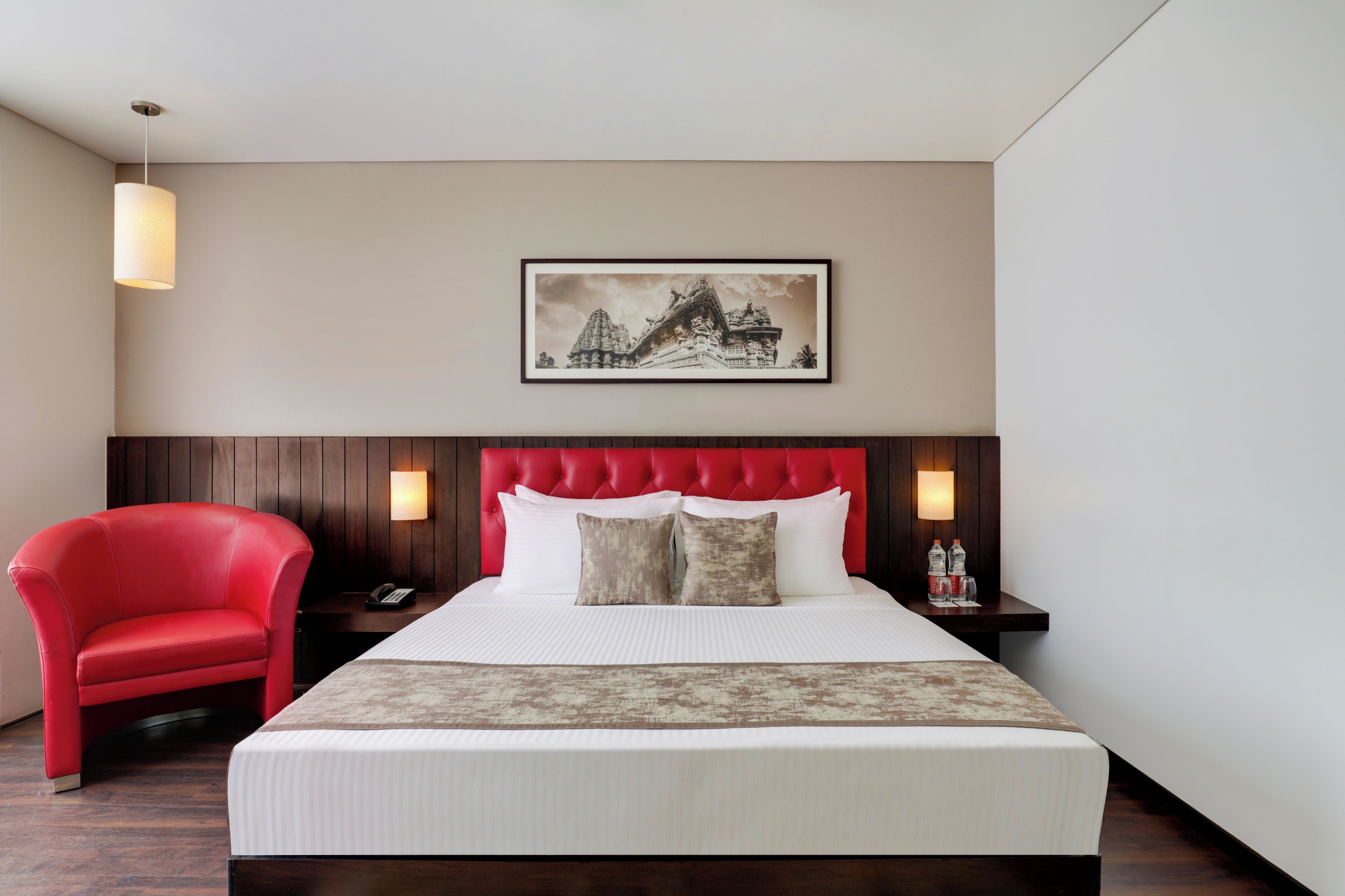 A hotel room with a bed, a red chair, a nightstand with a phone, a framed picture on the wall, and a wall-mounted lamp.