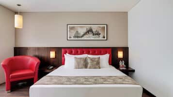 A hotel room with a bed, a red chair, a nightstand with a phone, a framed picture on the wall, and a wall-mounted lamp.