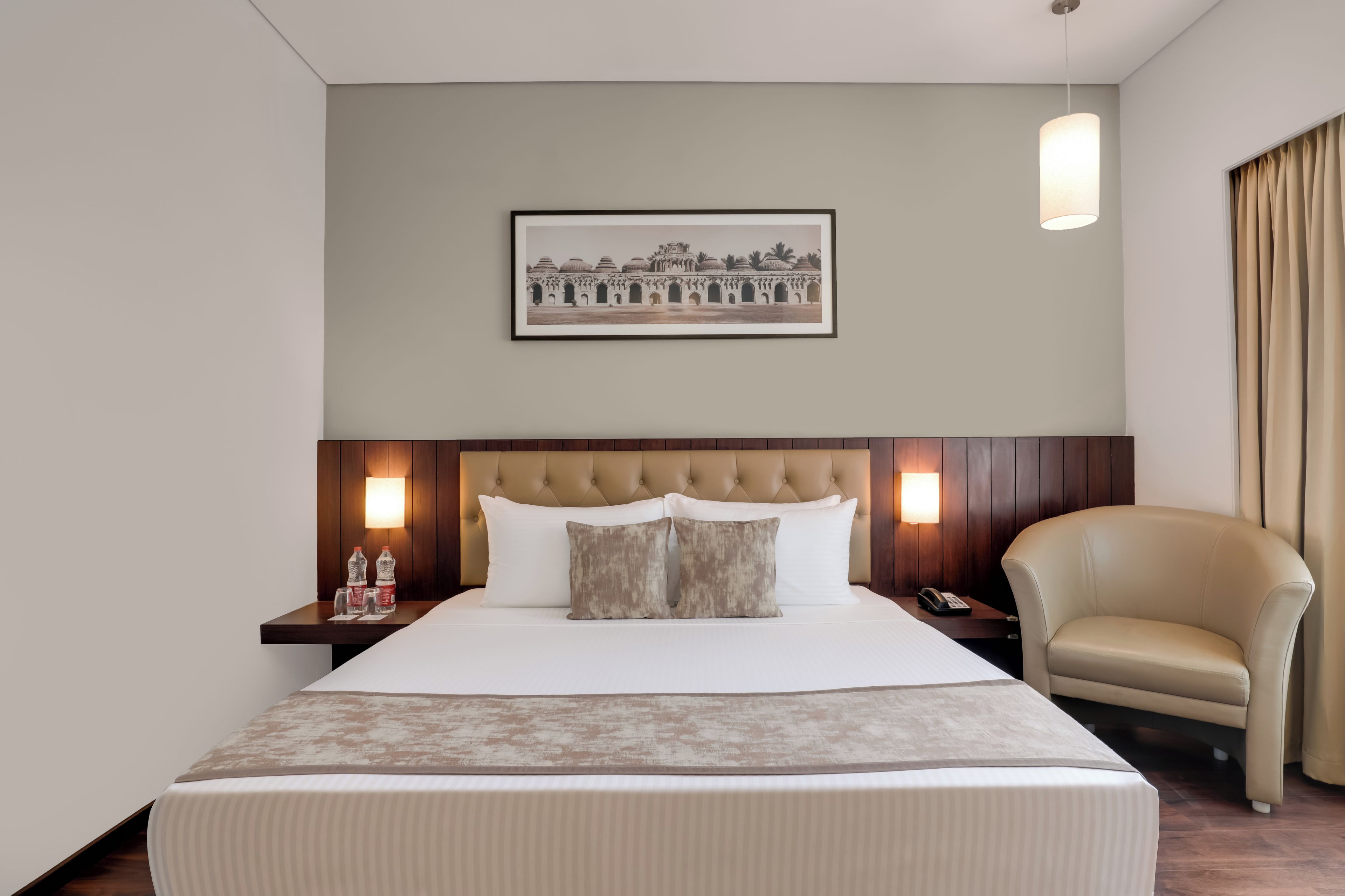 Une chambre d’hôtel avec un grand lit, une tête de lit dotée d’un luminaire, un tableau encadré au mur, une table de chevet avec une lampe et un fauteuil en cuir.