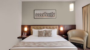 Una habitación de hotel con una cama grande, un cabecero con lámpara, un cuadro en la pared, una mesita de noche con lámpara y un sillón de cuero.