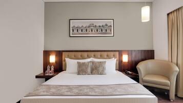 Une chambre d’hôtel avec un grand lit, une tête de lit dotée d’un luminaire, un tableau encadré au mur, une table de chevet avec une lampe et un fauteuil en cuir.