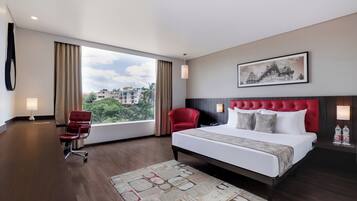 Ein Hotelzimmer mit einem großen Bett, einem roten Sessel, einem Schreibtisch und einem Fenster mit Blick auf die Stadt.
