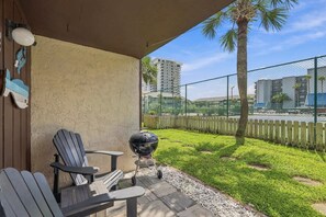 A patio with outdoor seating and a grill, overlooking a grassy area with a fence and buildings in the background. - Summer Place 127 | Sleeps 6 | Great Fall Break Vacation! (Panama City)