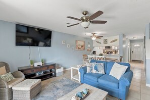 Un salon avec un canapé bleu, un téléviseur, un ventilateur de plafond et, au fond, un coin cuisine.