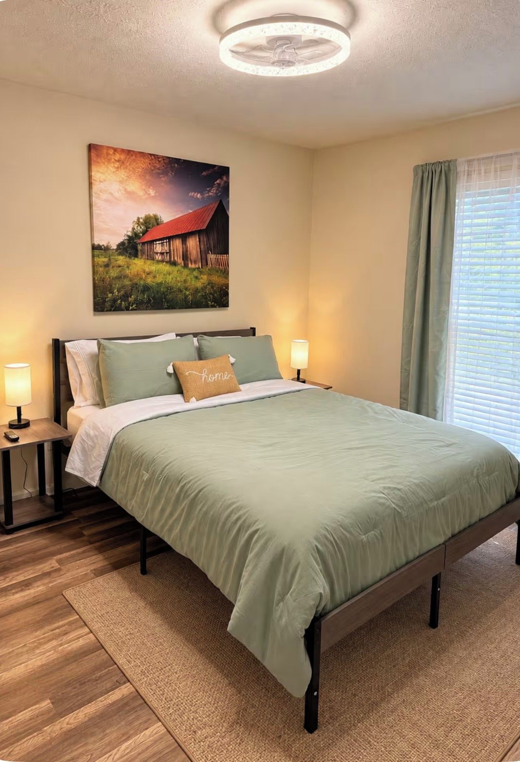A bedroom with a bed, two bedside tables, a nightstand, a wall-mounted lamp, a window with curtains, and a painting on the wall.
