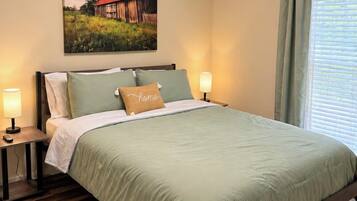 A bedroom with a bed, two bedside tables, a nightstand, a wall-mounted lamp, a window with curtains, and a painting on the wall.