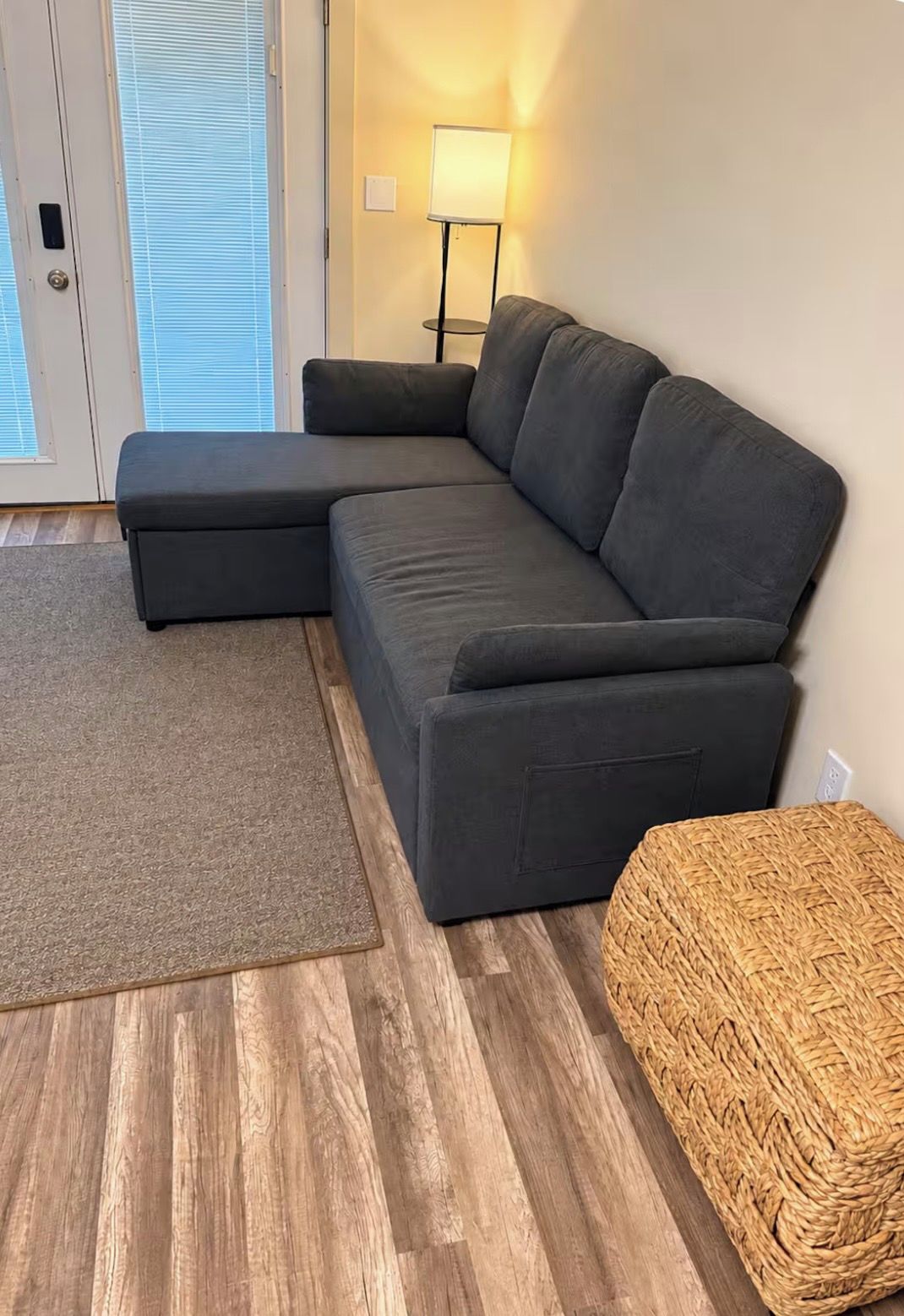 A sectional sofa in a living room with a woven ottoman and a lamp.