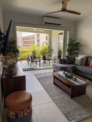 A modern living room with a sofa, coffee table, and a dining area with chairs. There is a ceiling fan and an air conditioning unit. - The Snapdragon Courtyard Garden (Montego Bay)