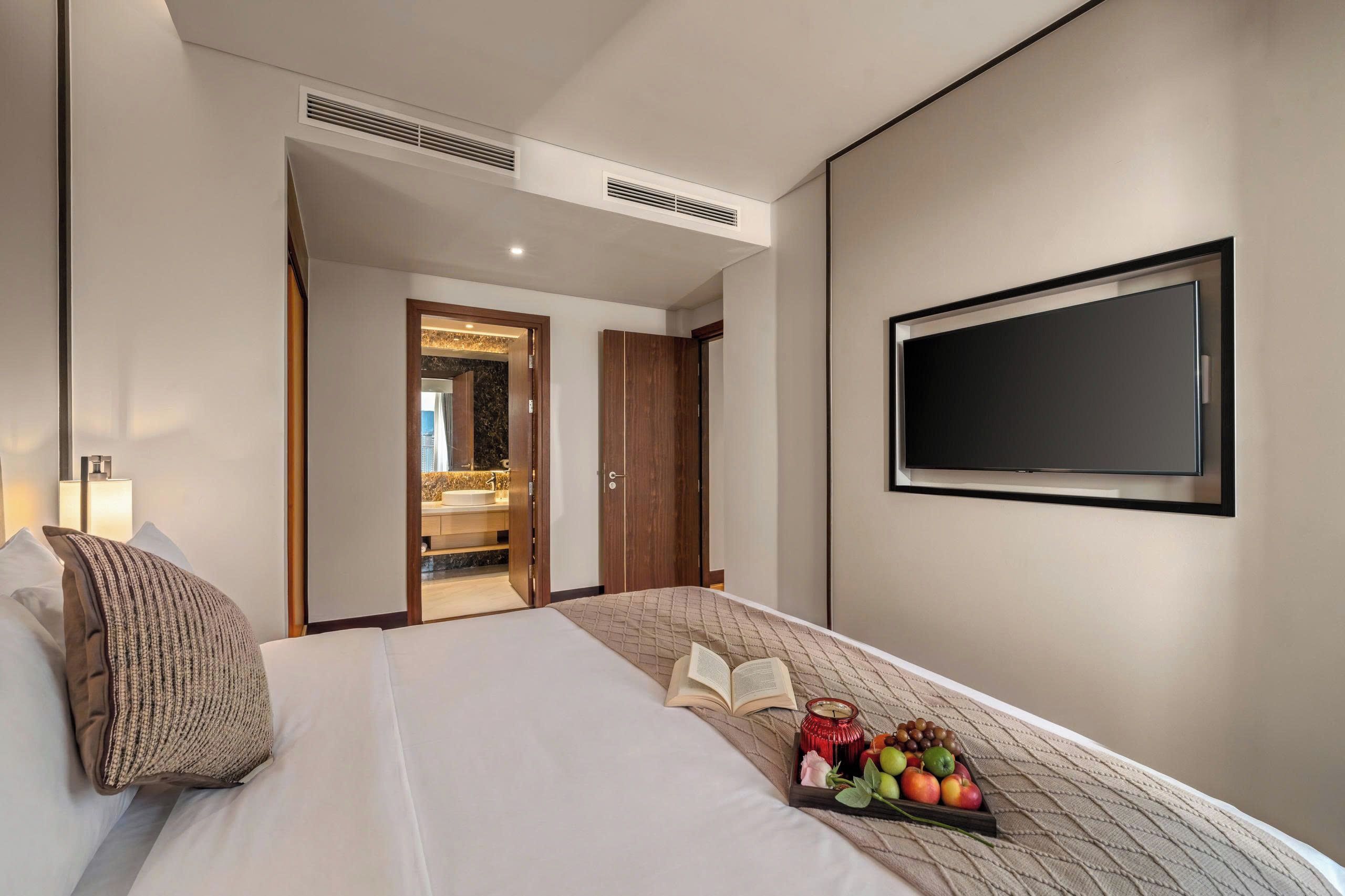 A hotel room with a bed, a flat-screen TV, a bedside table with a book and a candle, and a tray with fruits and a rose.