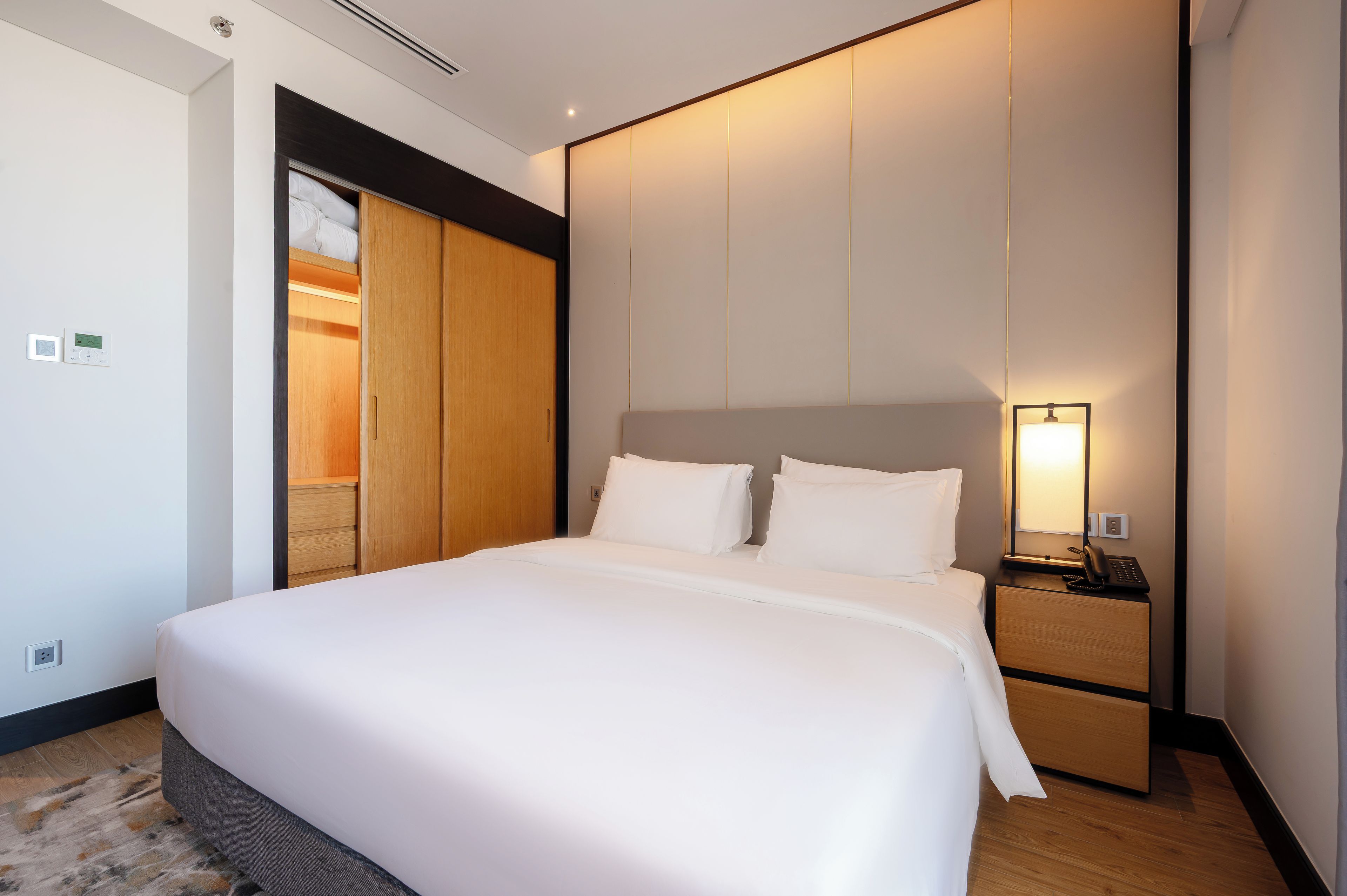 A hotel room with a large bed, a bedside table with a lamp, a wardrobe, and a thermostat on the wall.