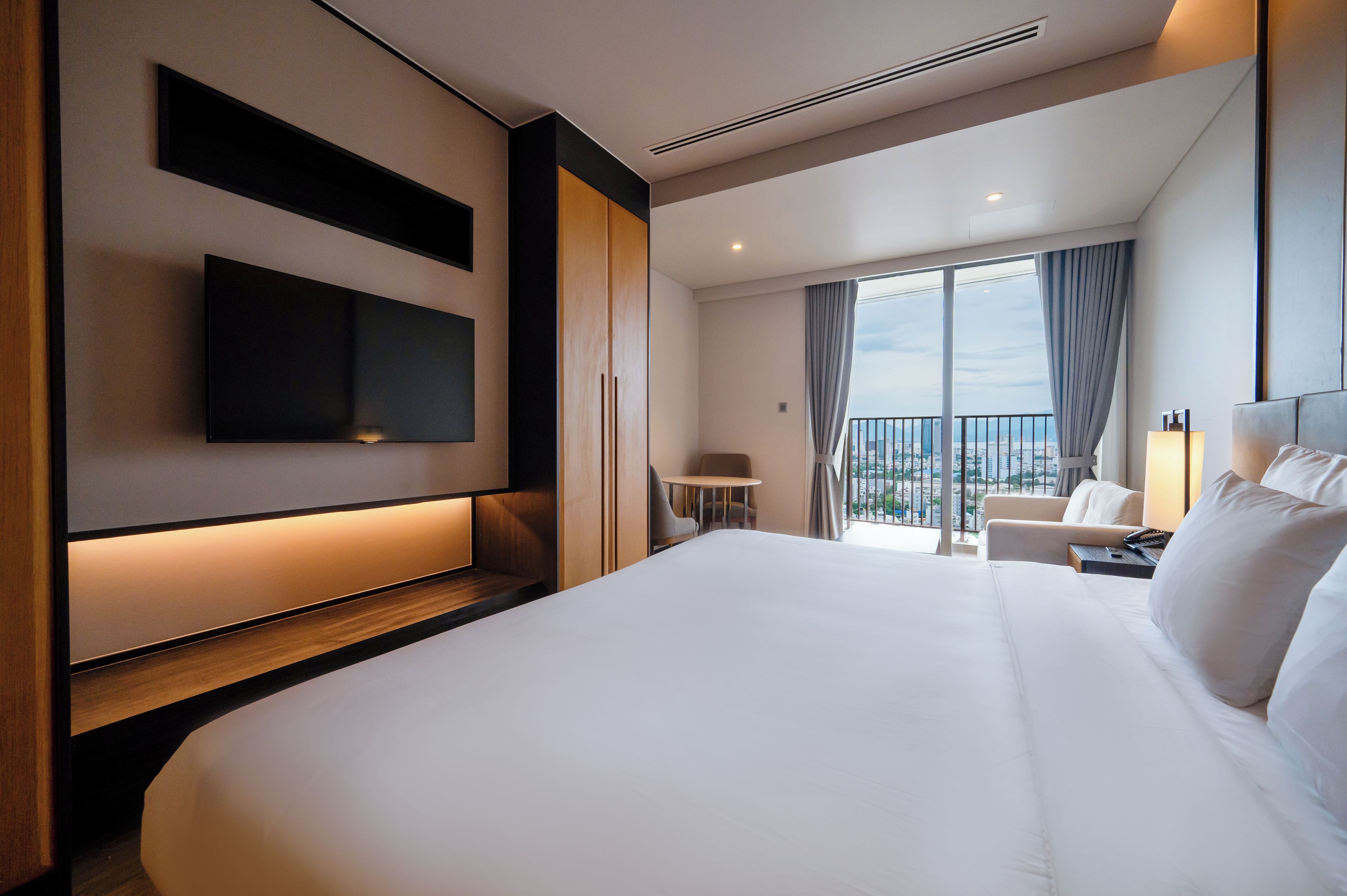 A hotel room with a large bed, a flat-screen TV mounted on the wall, a small table with a chair, and a view of the city through the window.