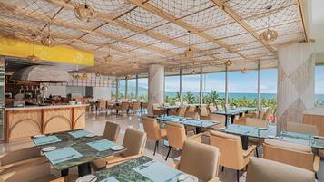 Een restaurant met uitzicht op de oceaan, met een hoog plafond en een netstructuur, houten tafels en stoelen.