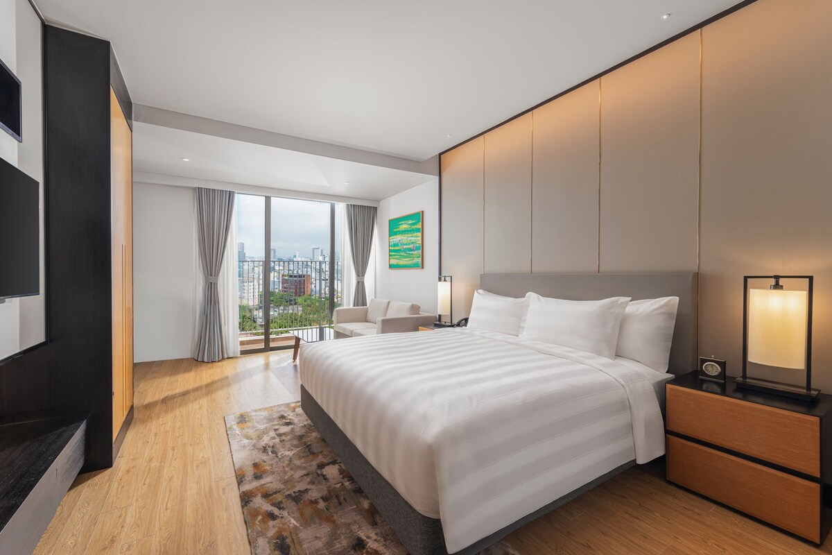A modern hotel room with a large bed, a desk, a TV, and a view of the city.