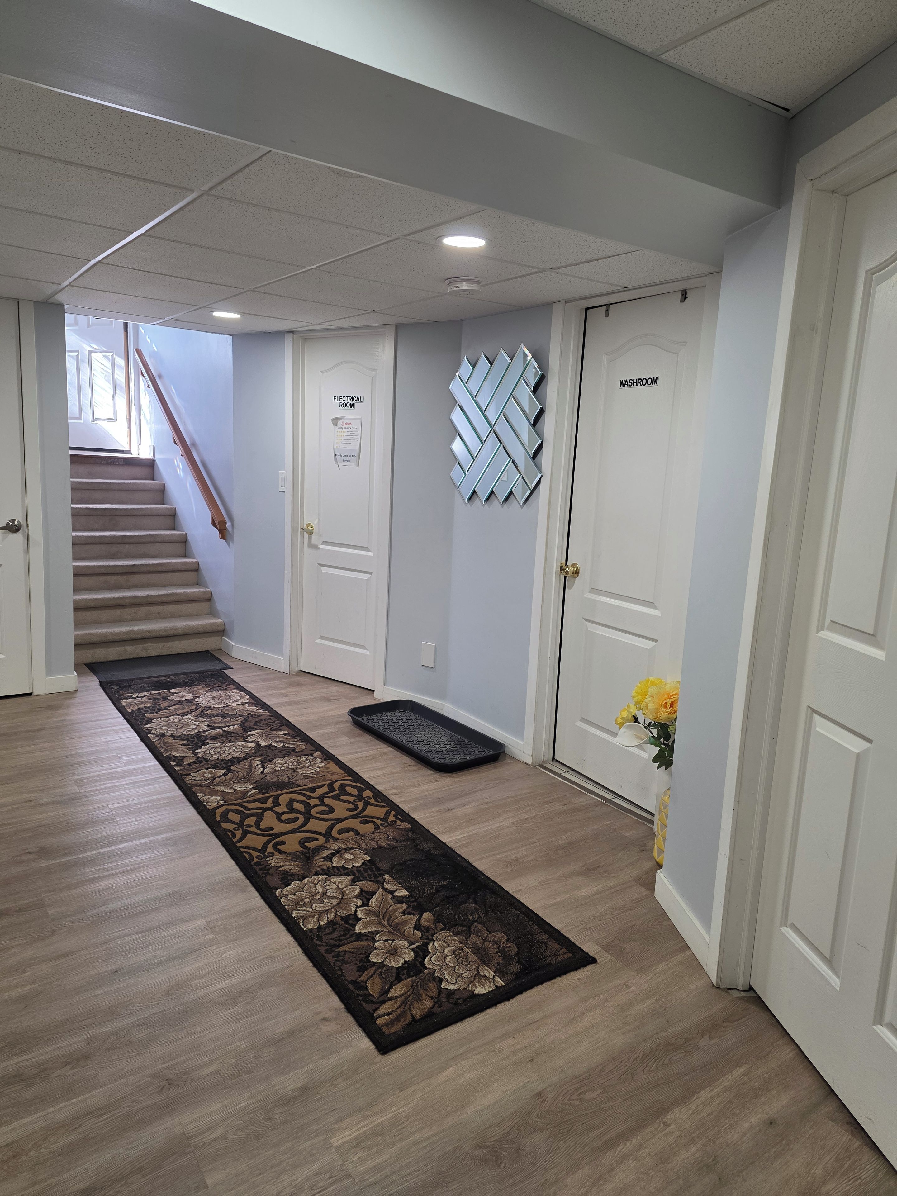 Un couloir avec un tapis, un escalier et des portes portant les inscriptions « Local électrique » et « Toilettes ».
