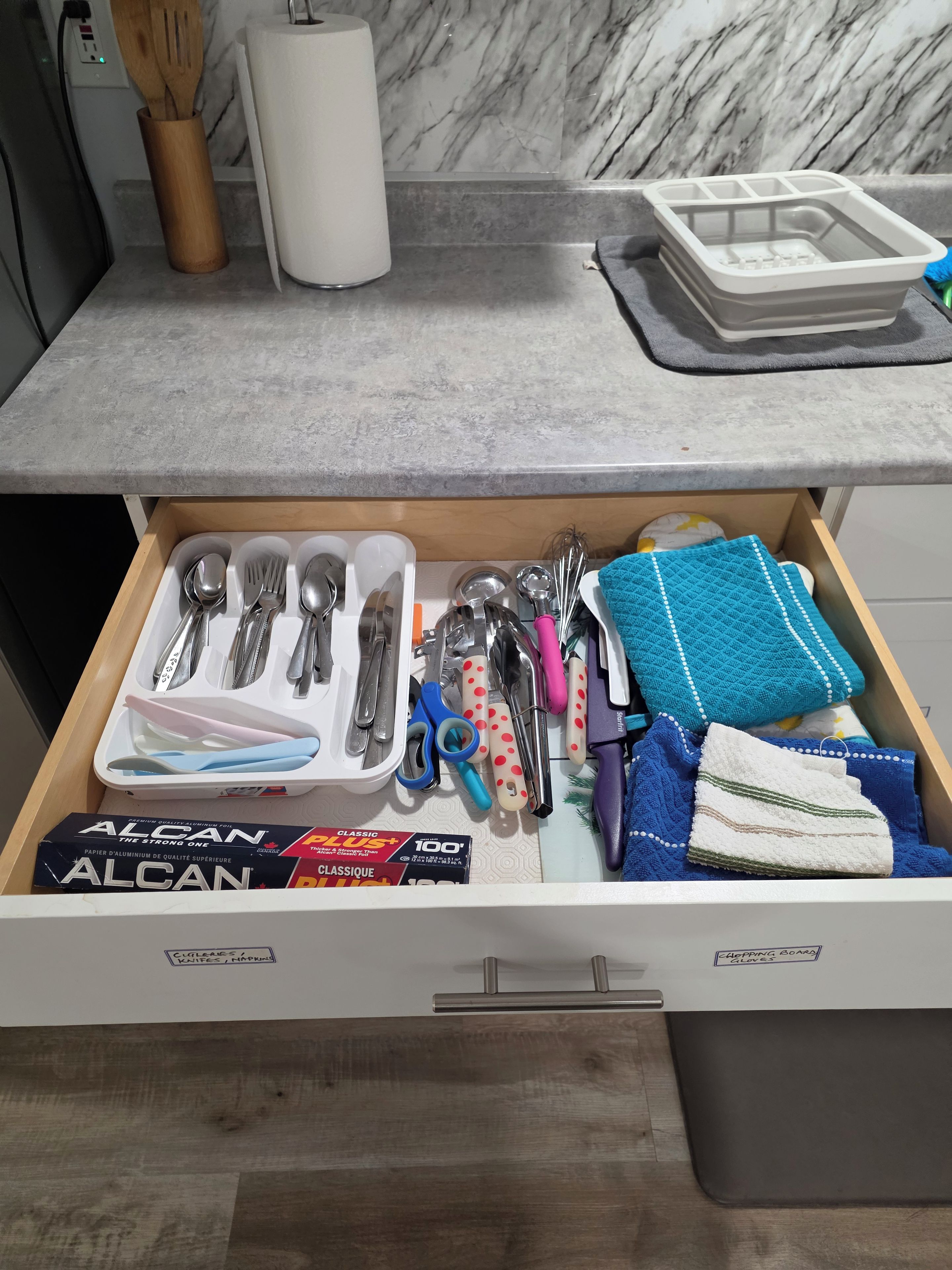 A kitchen drawer with utensils, a box of aluminum foil, and a dish rack.
