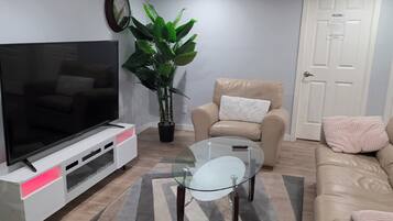 Una sala de estar con televisor de pantalla plana, una mesa de centro de vidrio, un sofá color beige, una planta en maceta y una alfombra.